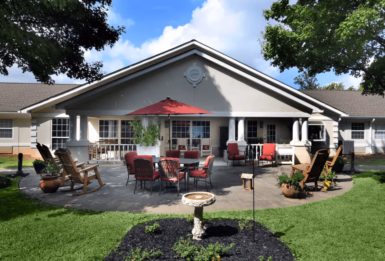 Outdoor patio area at The Waterford at Decatur featuring a circular concrete seating area with red cushioned chairs around a table with a red umbrella, wooden rocking chairs, potted plants, and a birdbath in the foreground, surrounded by green grass and trees under a partly cloudy sky.