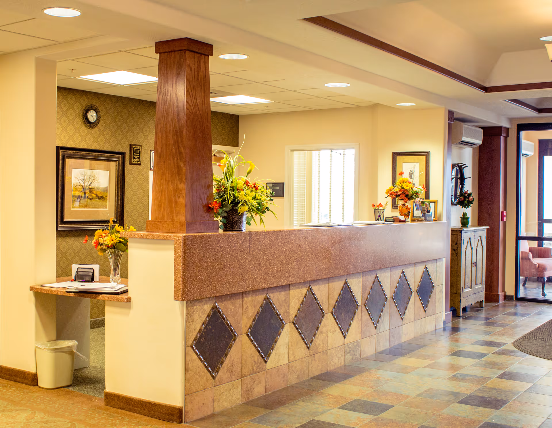 Reception desk in a senior living facility lobby with flowers, framed art, and a tiled floor.