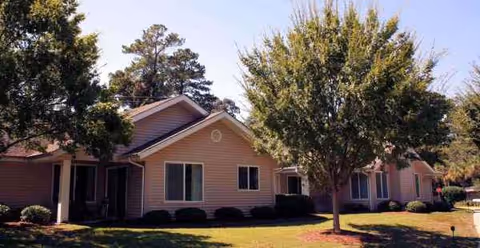 Single-story beige senior living building with windows, shrubs, and trees in a sunny yard.