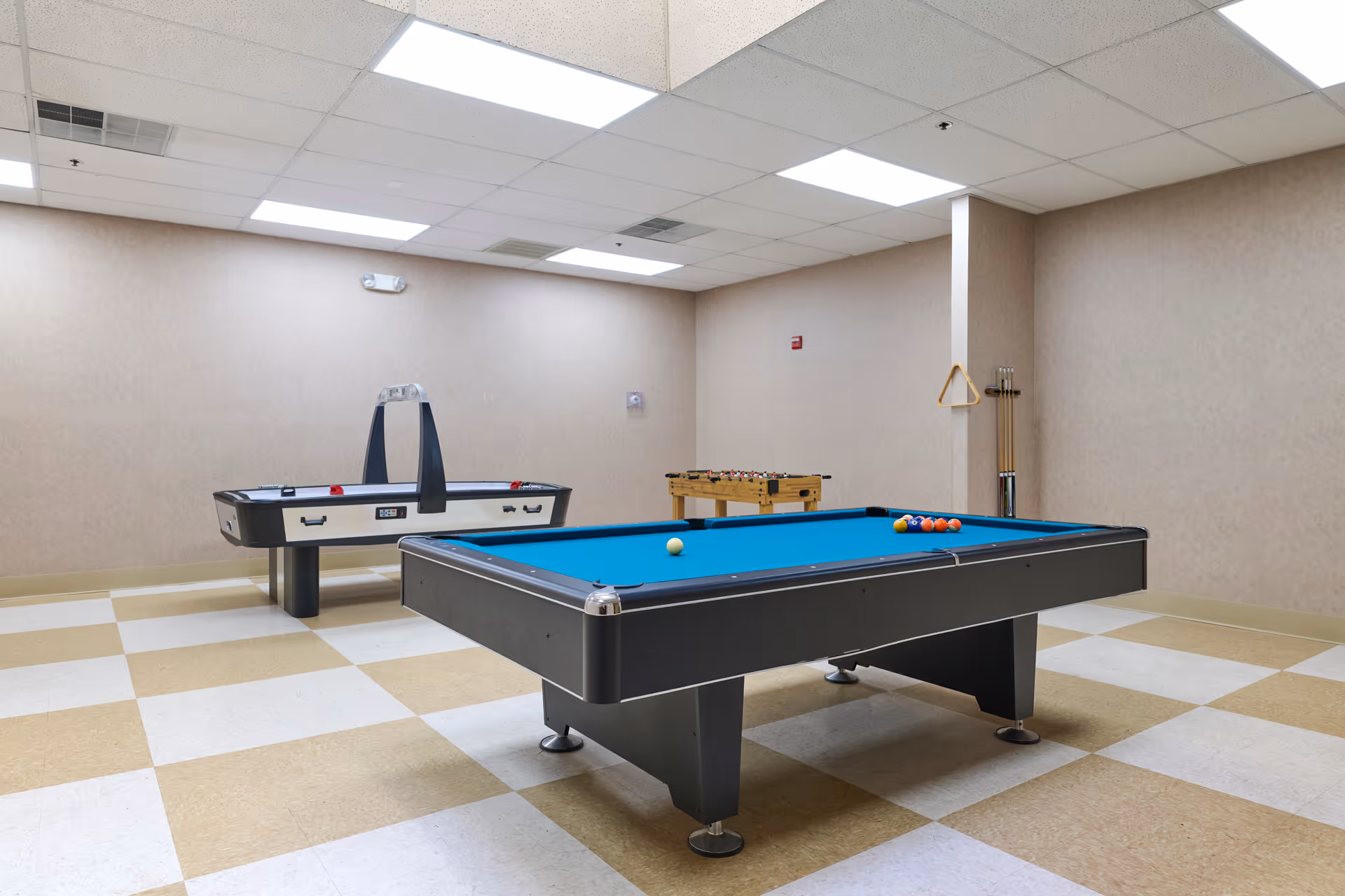Well-lit recreational room with a pool table, air hockey table, and foosball table on a checkered floor.
