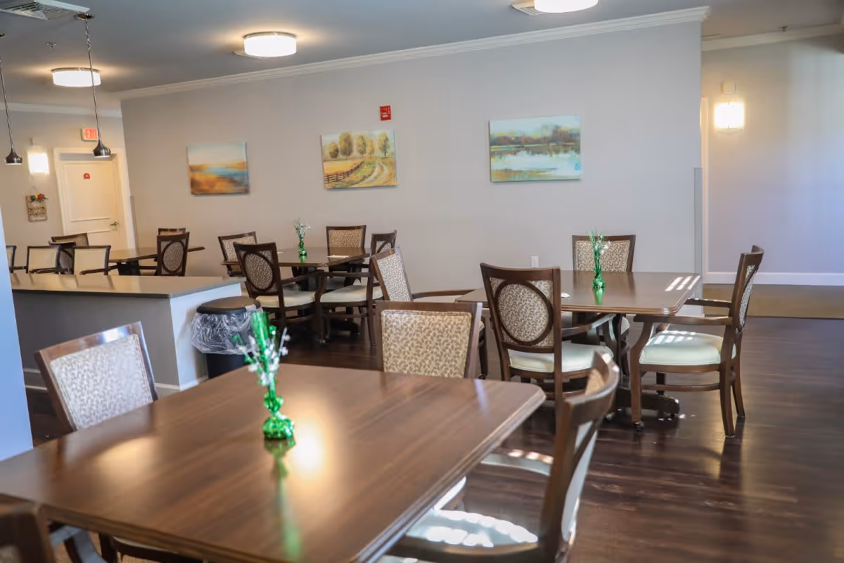 A dining room with several wooden tables and chairs arranged neatly. Each table has a small green vase with decorative white flowers. The room has light-colored walls adorned with landscape paintings and dark wood flooring. Ceiling lights and pendant lights illuminate the space.