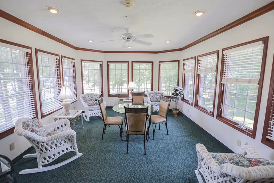Bright sunroom with many windows, wicker rocking chairs, floral cushions and a small round glass table with chairs.