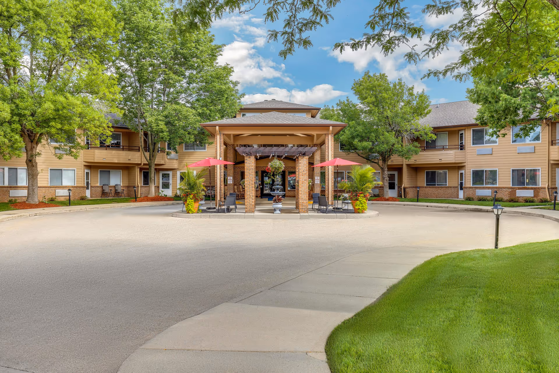 Front exterior view of Holiday Blair House senior living facility with a circular driveway, a covered entrance supported by brick columns, red umbrellas shading seating areas, green trees, and a well-maintained lawn under a partly cloudy sky.
