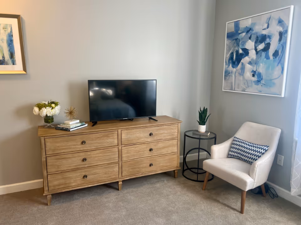 A wooden six-drawer dresser with a TV on top, an accent chair, side table, and wall art in a softly decorated room.