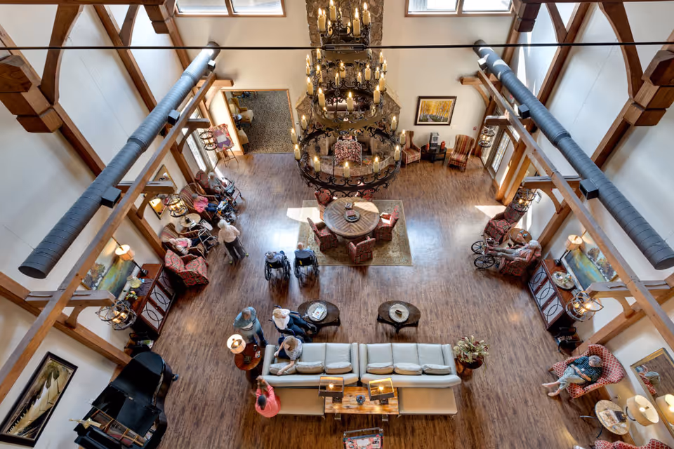 Aerial view of a senior living facility's large communal lounge with exposed wooden beams, a central chandelier, multiple seating areas, and residents gathered throughout.