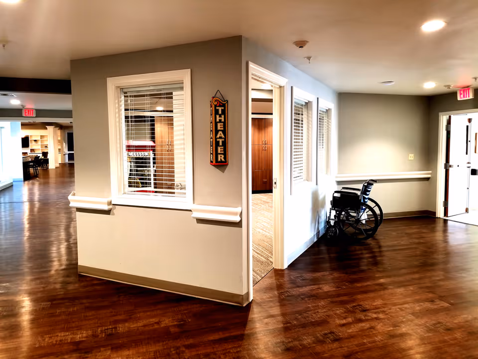 Well-lit interior hallway of a senior living facility with wood floors, a wheelchair by a doorway and a 'THEATER' sign on the wall.