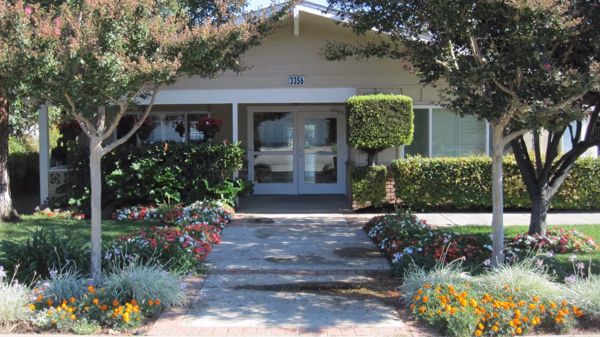 Front entrance of a single-story building with the number 3356 above the door, surrounded by well-maintained landscaping including trees, bushes, and colorful flowers along a concrete walkway.