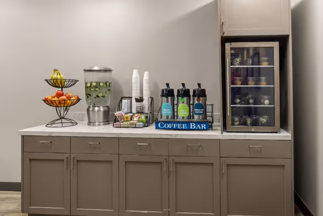 Countertop coffee bar with fruit tiers, a cucumber-infused water dispenser, disposable cups, coffee airpots, and a small refrigerated display on a cabinet.