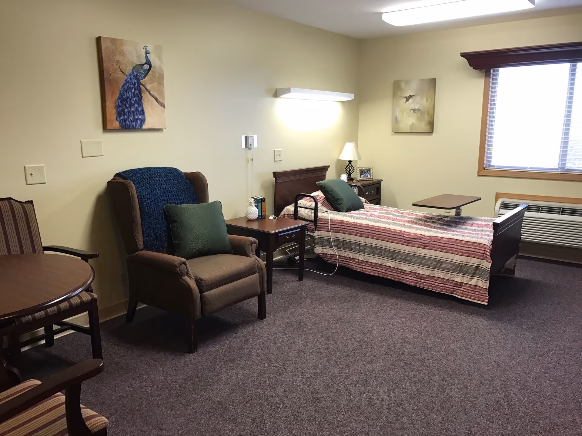 A senior living facility bedroom with a single bed covered in a striped quilt, a brown armchair with a green pillow and blue throw, a small wooden side table with a lamp and decorative items, a window with blinds, and two paintings on the beige walls.