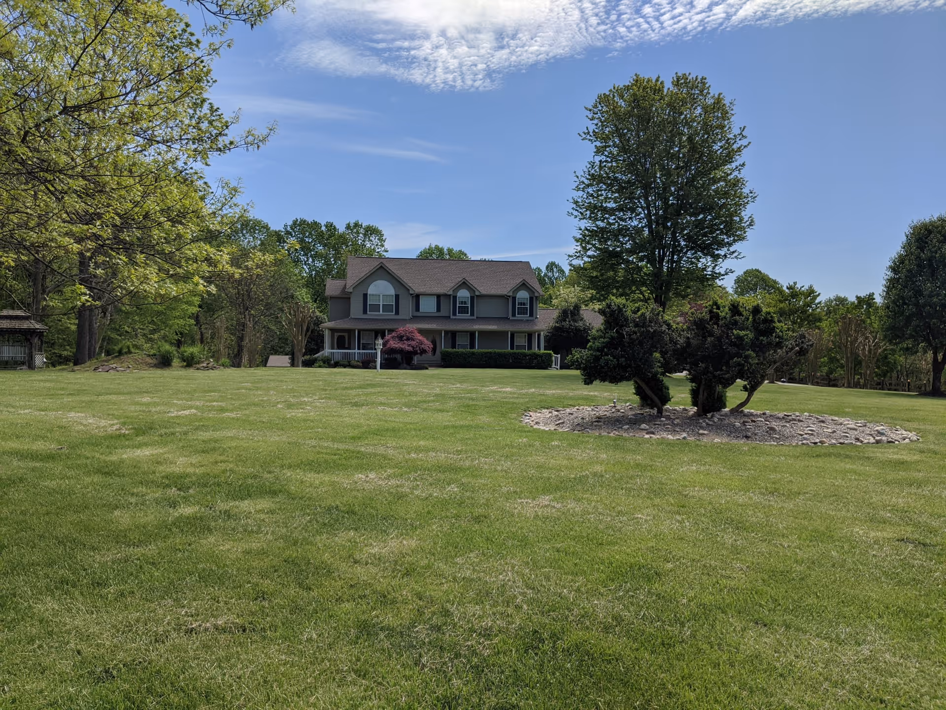 A large green lawn with a few trees and shrubs, leading up to a two-story house with a porch and multiple windows under a partly cloudy blue sky.