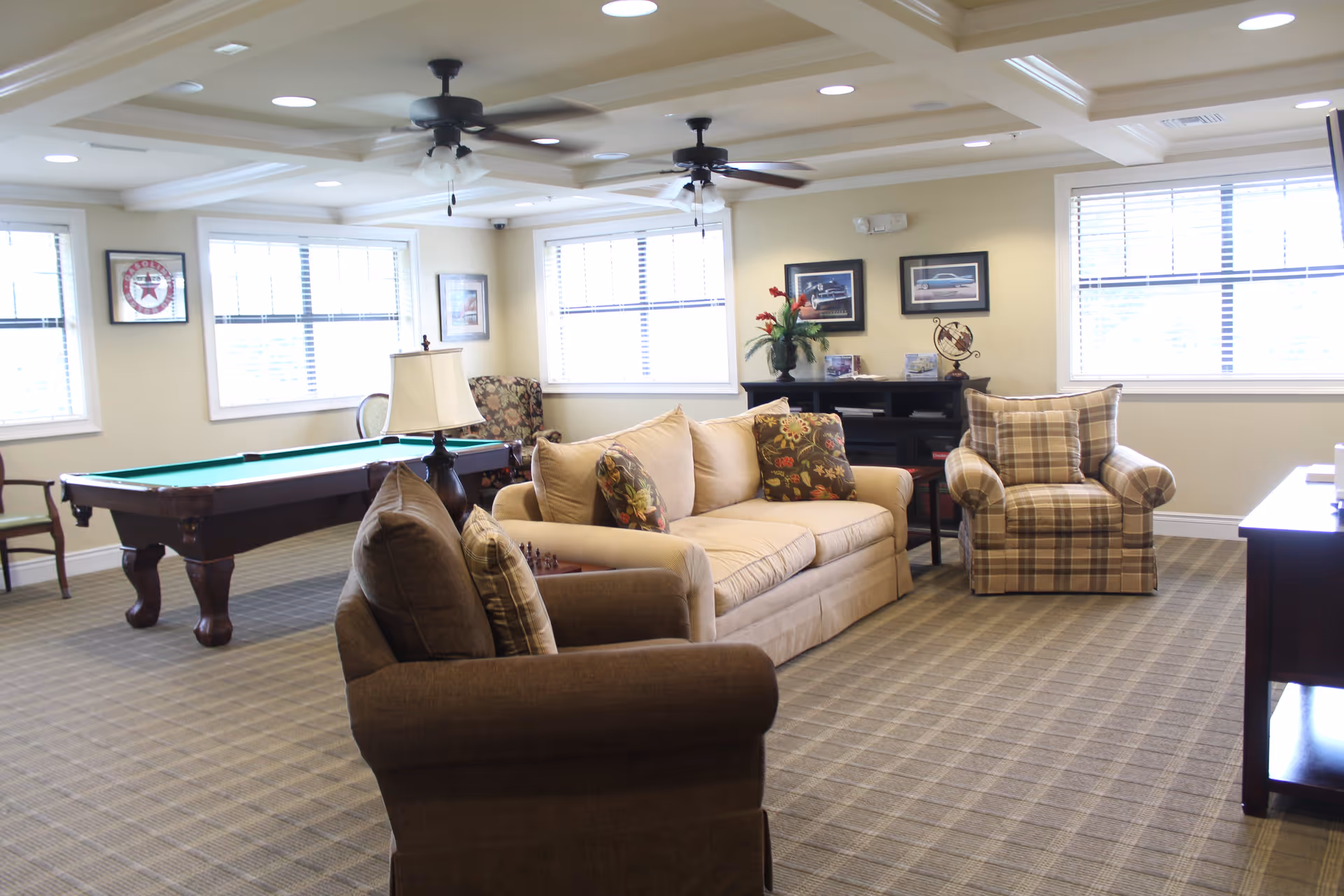 A bright and spacious living room area with a beige sofa, two armchairs, a pool table, and a lamp on a side table. The room has large windows with blinds, ceiling fans, framed pictures on the walls, and a patterned carpet.