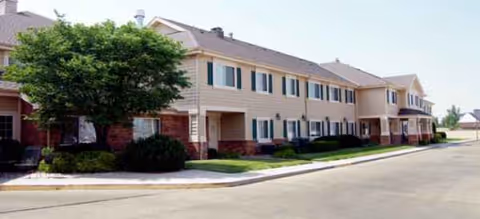 Two-story senior living building with beige siding, green shutters, and manicured lawn.