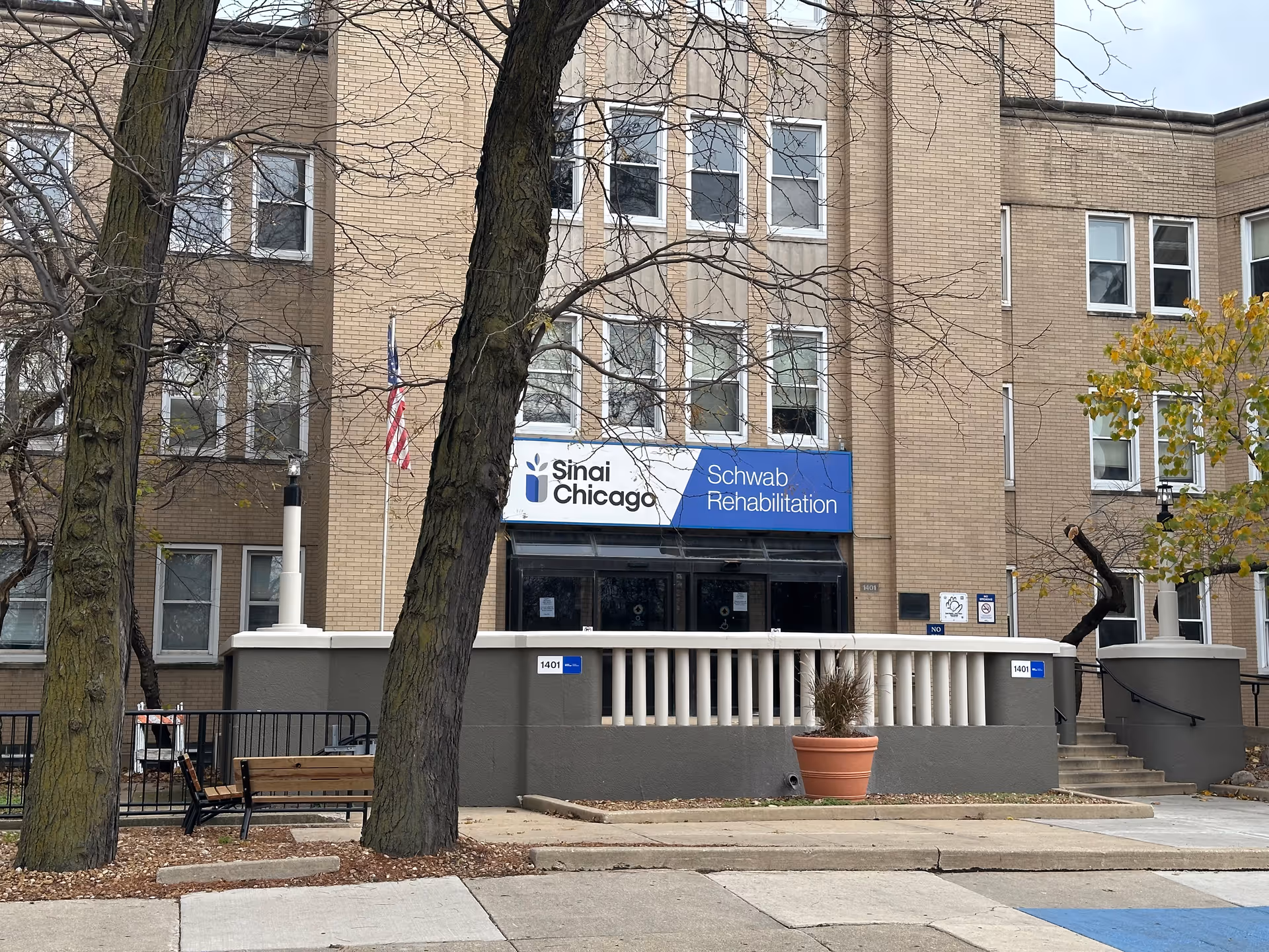 Front entrance of Sinai Chicago's Schwab Rehabilitation Center, a multi-story brick building with a blue sign, flagpole, and trees in front.