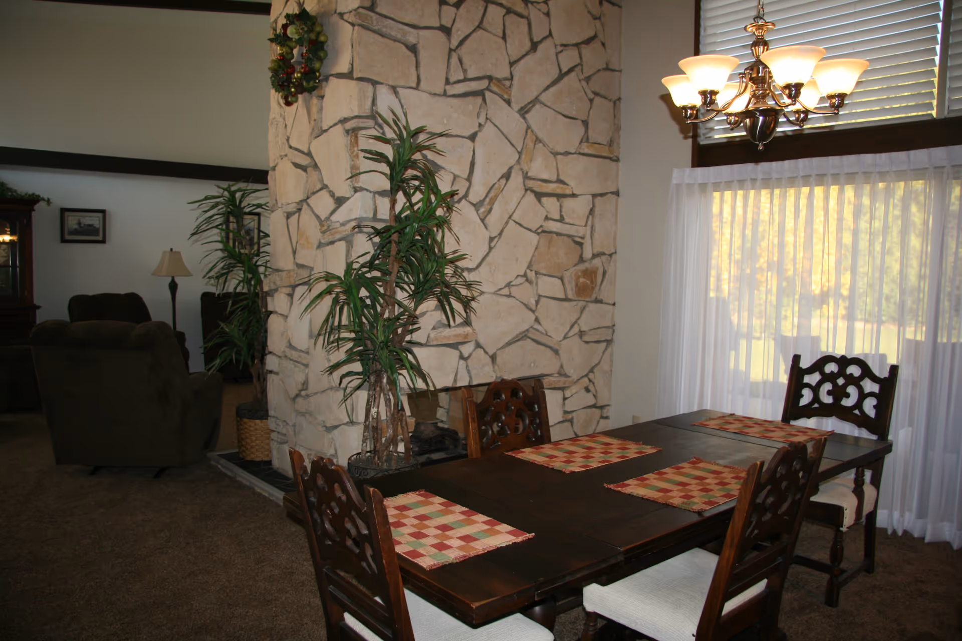 Interior view of a senior living facility dining area with a dark wooden table set with four chairs and checkered placemats. A large stone fireplace with green plants on either side separates the dining area from a living room with dark upholstered chairs and a floor lamp. A chandelier hangs above the dining table, and large windows with sheer white curtains allow natural light to brighten the space.