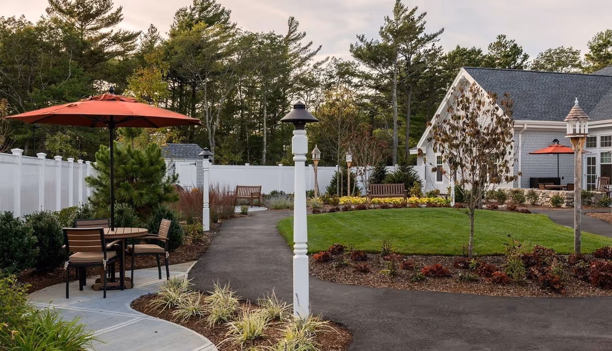 Well-maintained outdoor courtyard with paved walking paths, lamp posts, benches, landscaped lawn, and a patio table with a red umbrella near a building.