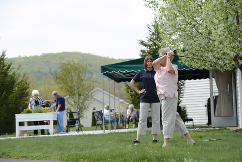 Two elderly women standing on a grassy lawn near a blooming tree, one woman is swinging a golf club while the other watches. In the background, other seniors are engaged in gardening and sitting under a green canopy with a building and trees visible.