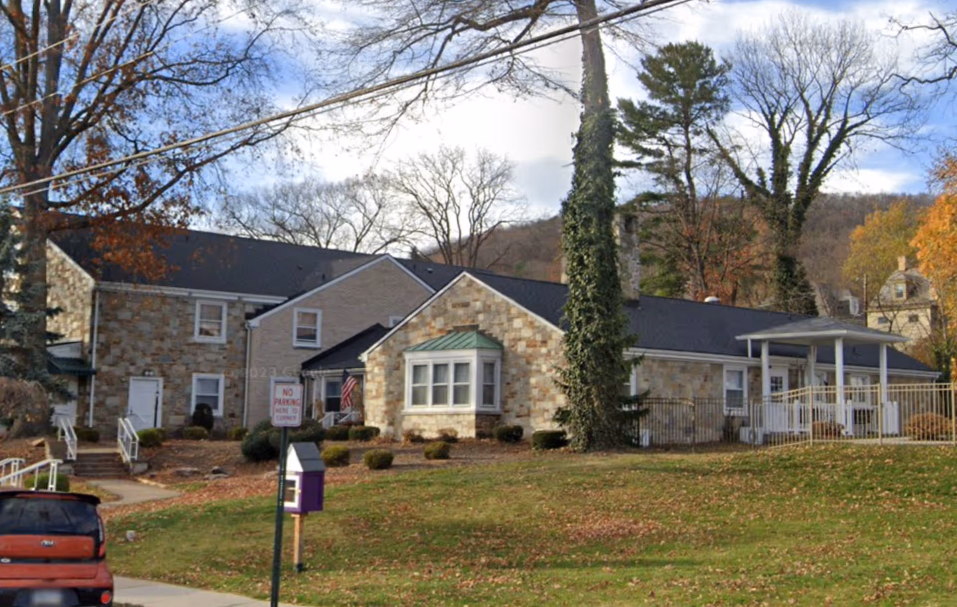 Exterior view of a stone and brick building with a black roof, surrounded by trees with autumn leaves. There is a small porch with white railings and an American flag near the entrance. A red car is parked on the street in front of the building, and a signpost with a 'No Parking' sign is visible on the grassy area.
