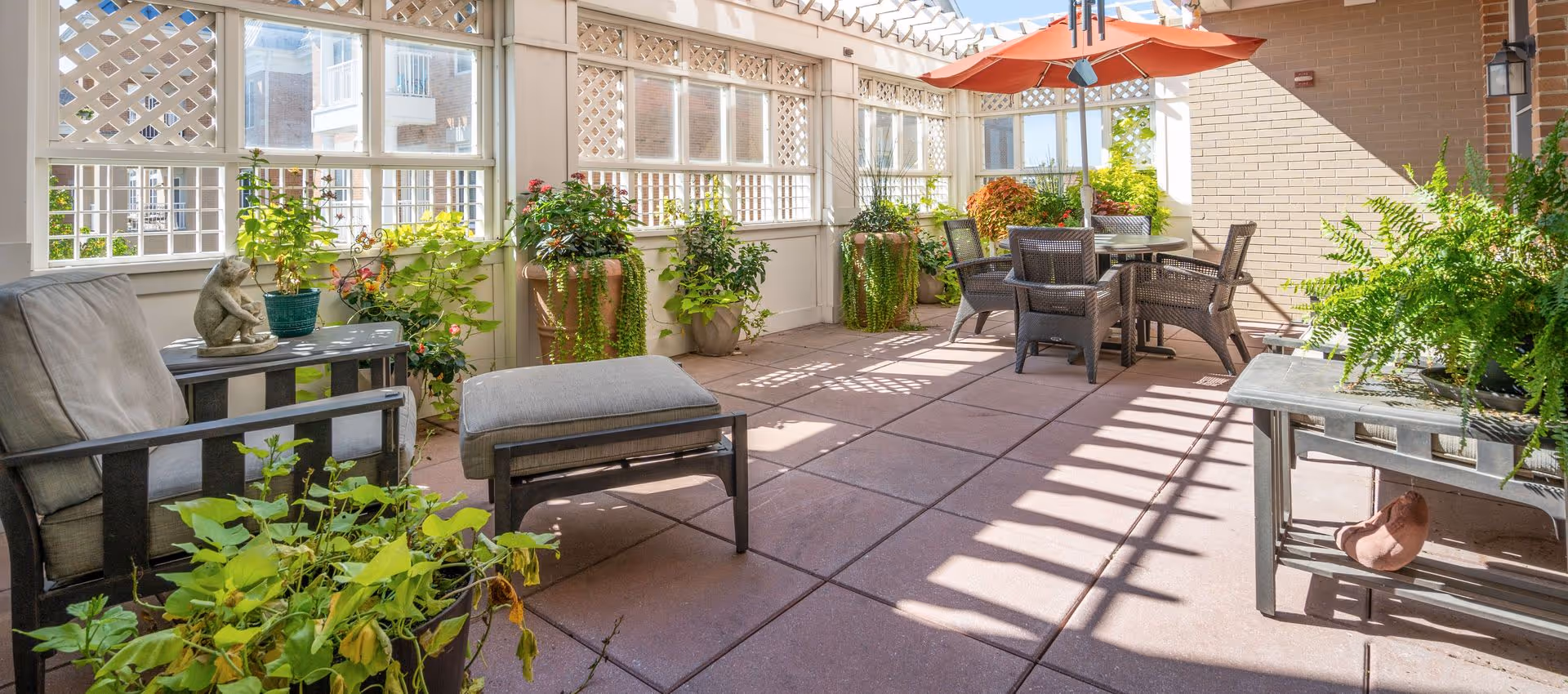 A bright and sunny enclosed patio area with cushioned chairs, a table with an orange umbrella, and various potted plants along the perimeter. The space has large windows with lattice detailing and a tiled floor.