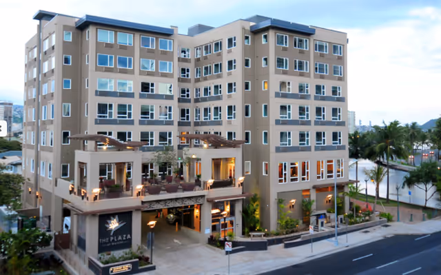Exterior view of The Plaza at Waikiki, a multi-story senior living facility with a modern design, balconies, and an entrance area with outdoor seating. The building is surrounded by palm trees and a street with a sidewalk.