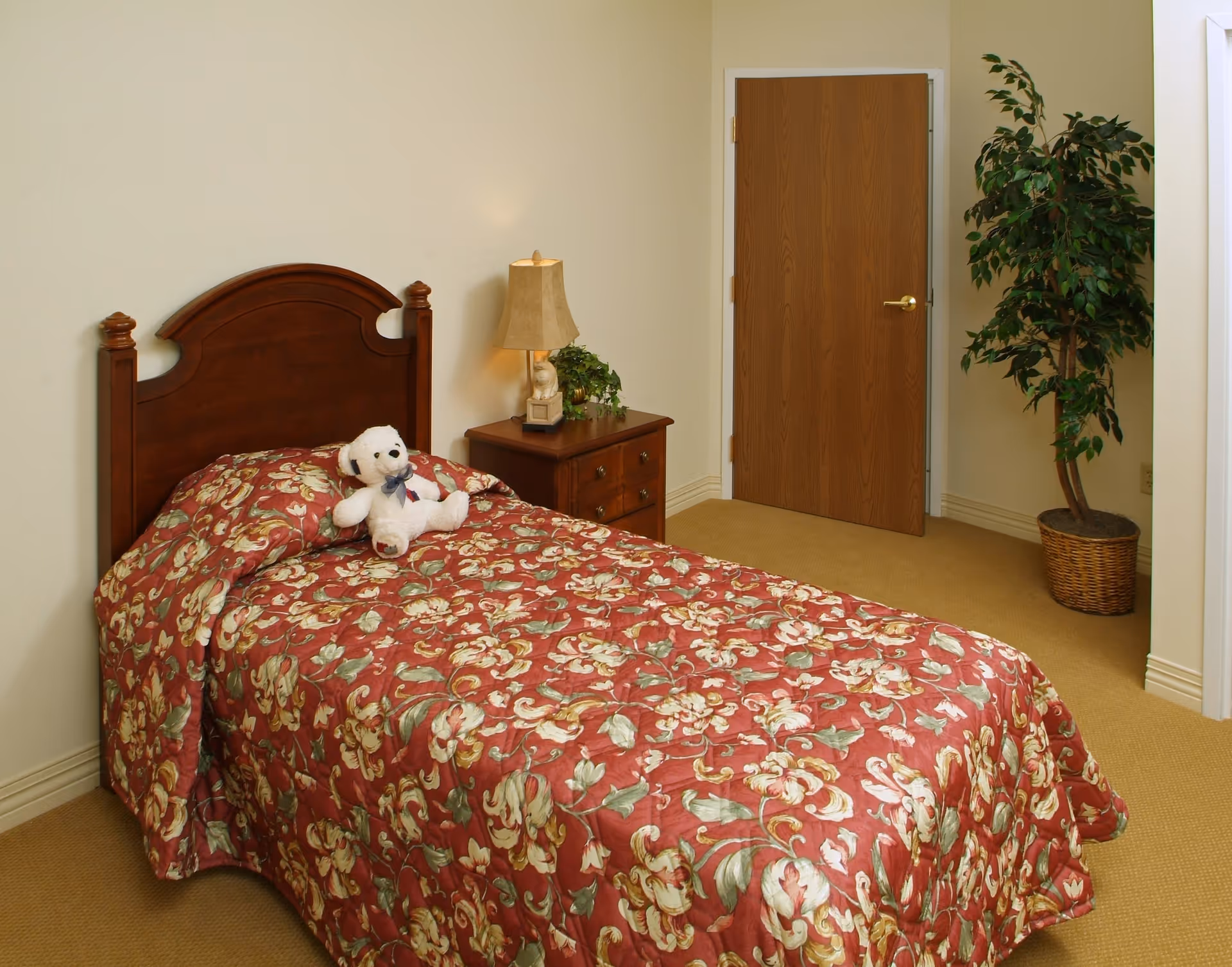 A bedroom with a single wooden bed covered with a red floral bedspread and a white teddy bear on top. Next to the bed is a wooden nightstand with a lamp and a small plant. There is a closed wooden door and a potted plant in the corner of the room.