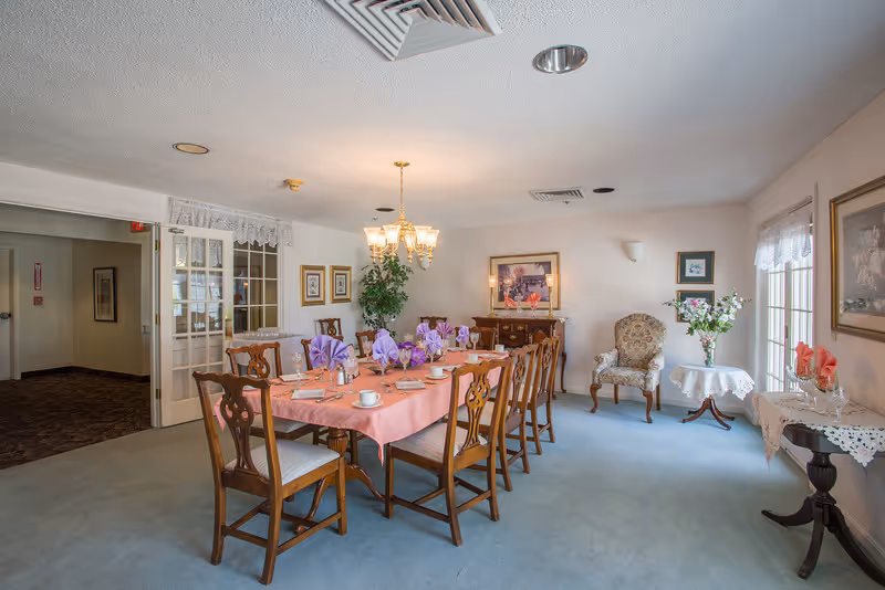 A dining room with a long wooden table covered with a pink tablecloth and set with cups, plates, and purple napkins folded in a decorative manner. The room has wooden chairs around the table, a chandelier hanging from the ceiling, framed artwork on the walls, a floral armchair, and two small tables with lace tablecloths and flower arrangements. There is a window with lace curtains letting in natural light.