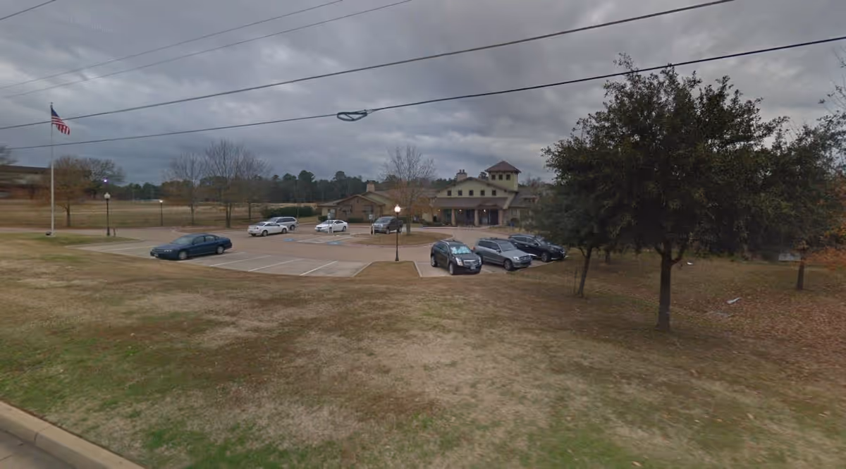 Exterior view of Hawkins Creek Assisted Living and Memory Care facility showing a parking lot with several cars, a large building in the background, trees, and an American flag on a flagpole under a cloudy sky.