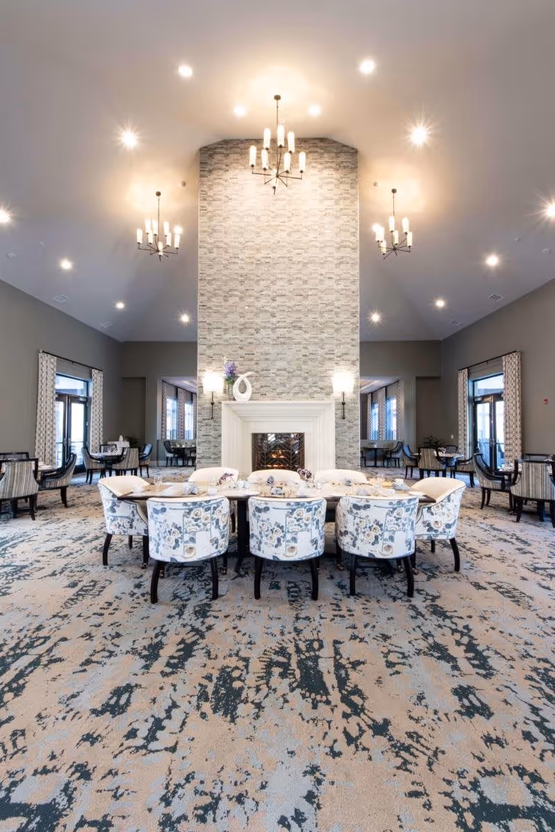 A spacious and elegant dining area with a large rectangular table surrounded by floral upholstered chairs. The room features a tall stone fireplace in the center with decorative vases on the mantel. Multiple chandeliers hang from the high ceiling, and large windows with patterned curtains allow natural light to fill the space. Additional smaller tables and chairs are visible in the background.