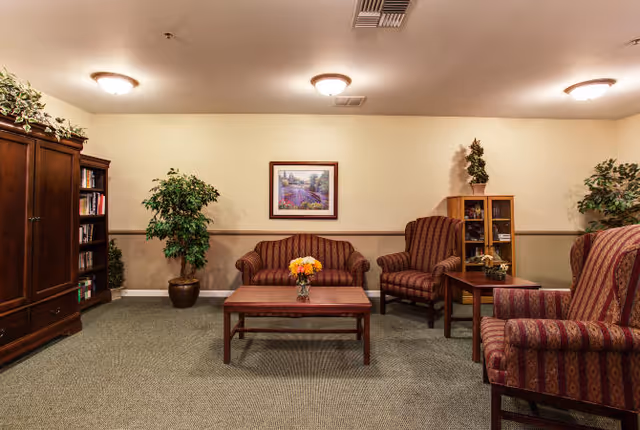 A cozy living room area with a striped sofa and two matching armchairs arranged around a wooden coffee table with a vase of flowers. There are potted plants on either side of the room, a wooden cabinet with books on the left, and a small display cabinet with decorative items on the right. A framed landscape painting hangs on the wall above the sofa. The room has beige walls, carpeted flooring, and ceiling lights.