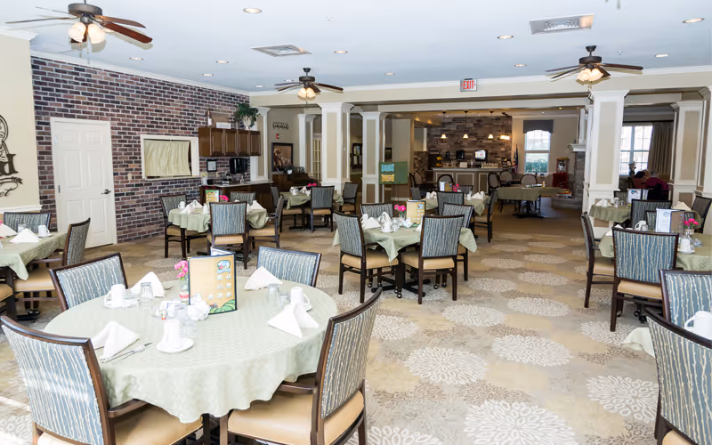 A spacious dining room with multiple round tables covered with light green tablecloths, each set with white napkins, cups, and small flower vases. The room features ceiling fans, a brick accent wall, and a carpet with a floral pattern. There is a kitchen area visible in the background with wooden cabinets and a counter.