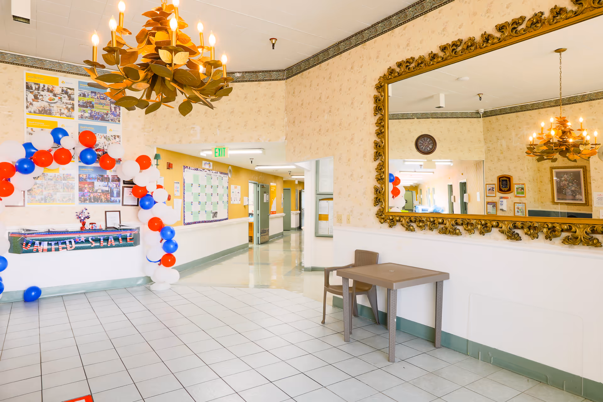 Interior hallway of a senior living facility decorated with a red, white, and blue balloon arch and a banner that reads 'United States'. There is a large ornate gold-framed mirror on the right wall reflecting part of the hallway and decorations. A small table and chair are positioned beneath the mirror. The hallway is well-lit with a decorative chandelier hanging from the ceiling.