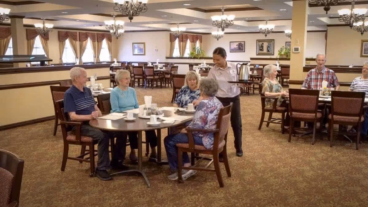 A group of elderly people sitting around tables in a spacious dining room at University Village Retirement Community. A staff member is serving coffee to one of the tables. The room is well-lit with chandeliers and has large windows with curtains.