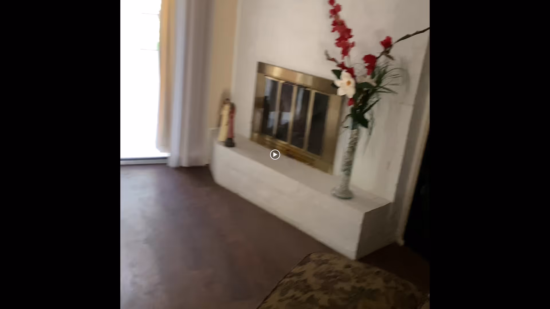 Interior view of a living room area featuring a fireplace with a brass frame, a vase with red and white flowers on the hearth, a small decorative statue, and a sliding glass door with white curtains allowing natural light in.