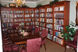 Community library-style room with wall-to-wall wooden bookshelves, a large polished table surrounded by upholstered chairs, potted plants, and a chandelier.