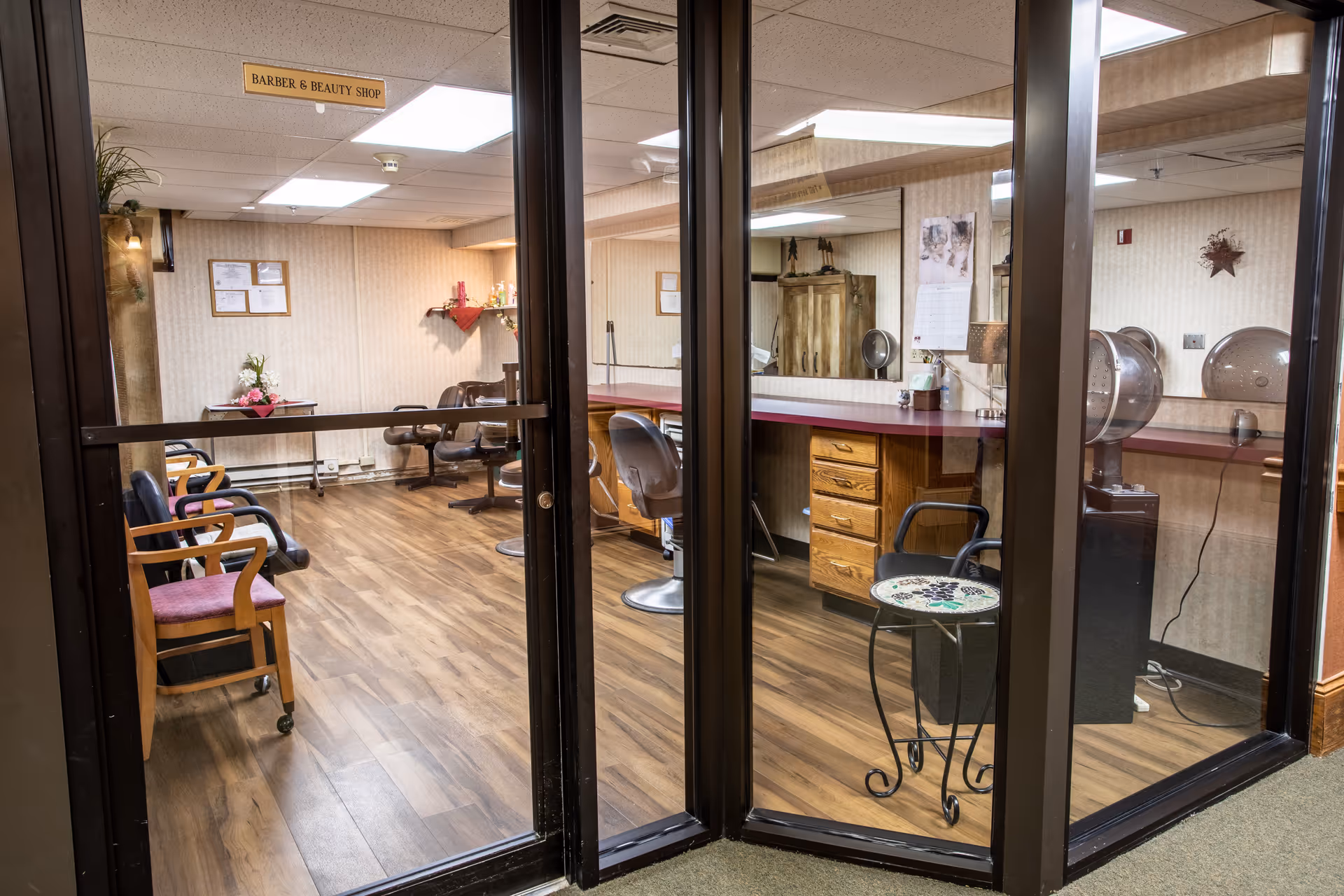 Interior view of a barber and beauty shop in a senior living facility, featuring wooden flooring, several chairs including salon styling chairs, a counter with drawers, hair drying stations, and a small table with a plant. The room is enclosed by glass walls and a door with a sign above reading 'Barber & Beauty Shop'.