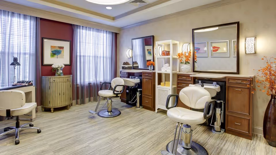 A well-lit salon area with two white salon chairs in front of wooden cabinets and sinks for hair washing. There is a white shelving unit with towels and decorative items, a small cabinet with a vase of flowers, and large windows with sheer curtains letting in natural light. The walls are decorated with framed artwork and a large mirror.