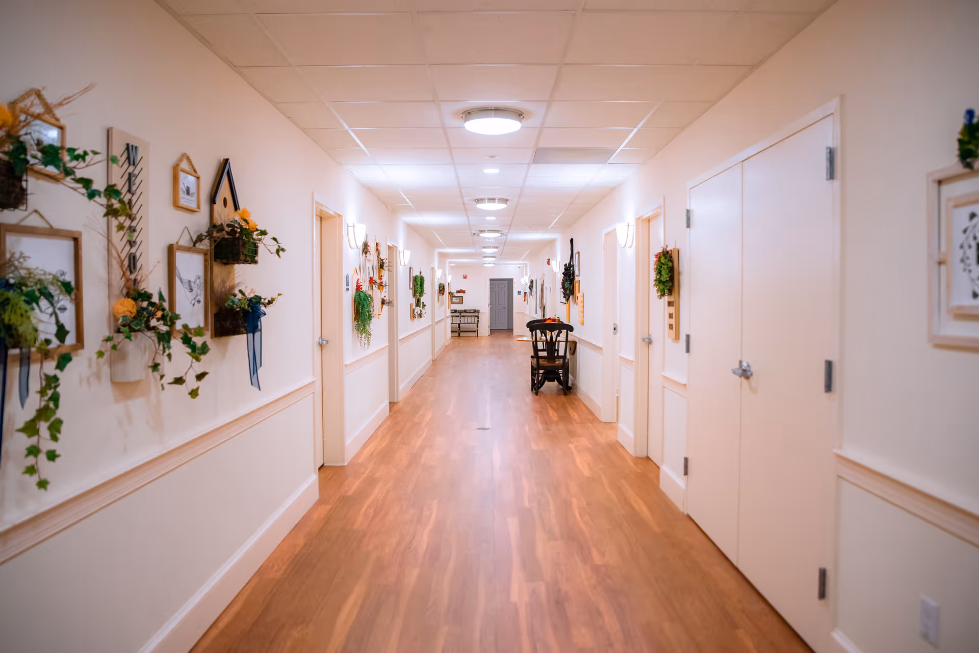 A long, well-lit hallway in a senior living facility with wooden flooring and white walls decorated with framed pictures and hanging plants. Several closed doors line both sides of the hallway, and a chair is positioned along the right wall.