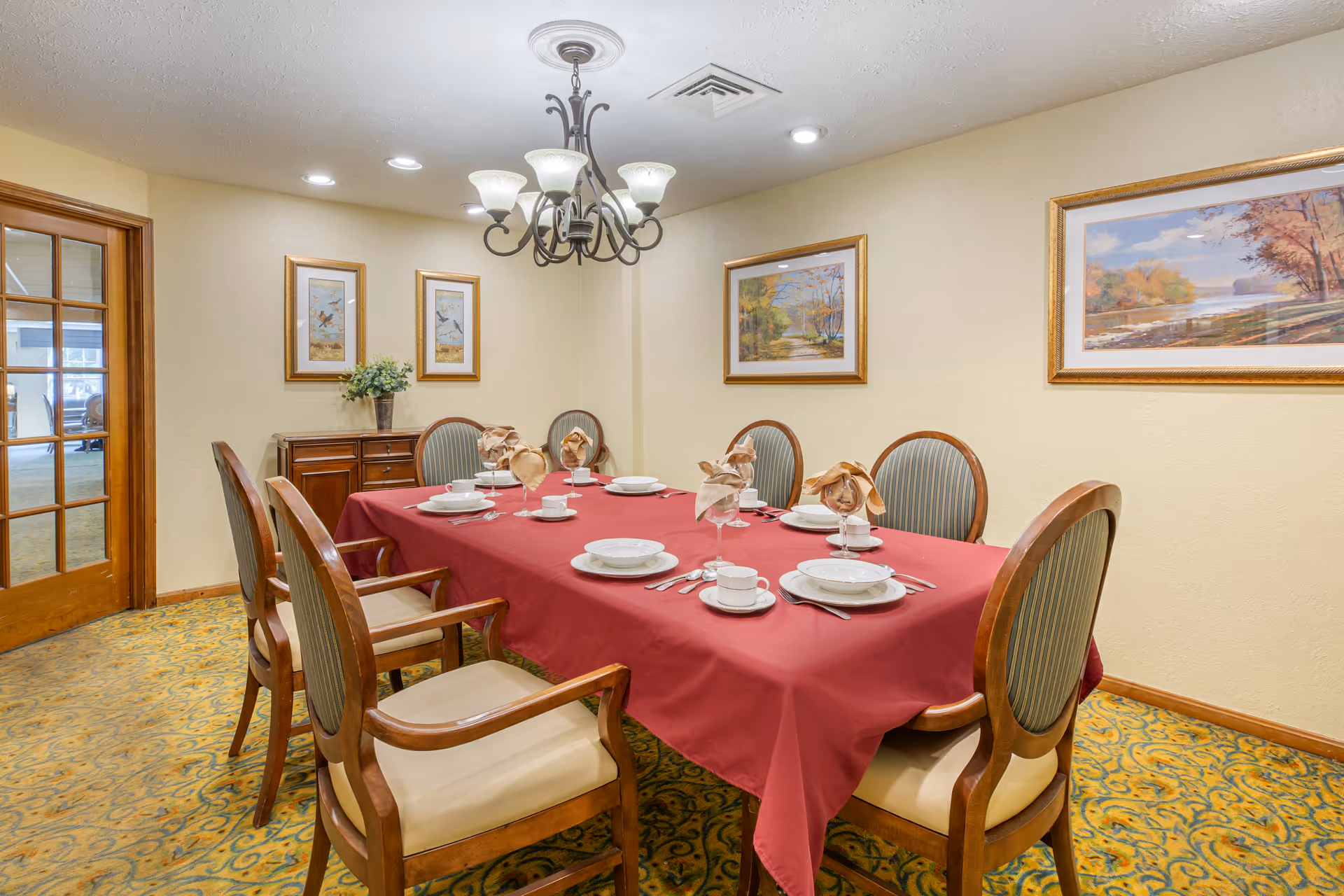 A dining room with a rectangular table covered with a red tablecloth set for six people. The table settings include white plates, bowls, cups, silverware, and glasses with folded beige napkins. The room has six wooden chairs with cushioned seats and backs. The walls are decorated with framed landscape paintings and two smaller framed pictures. A chandelier with six lights hangs above the table, and there is a wooden sideboard against the wall. The floor is carpeted with a patterned design, and a wooden door with glass panels is visible on the left side.