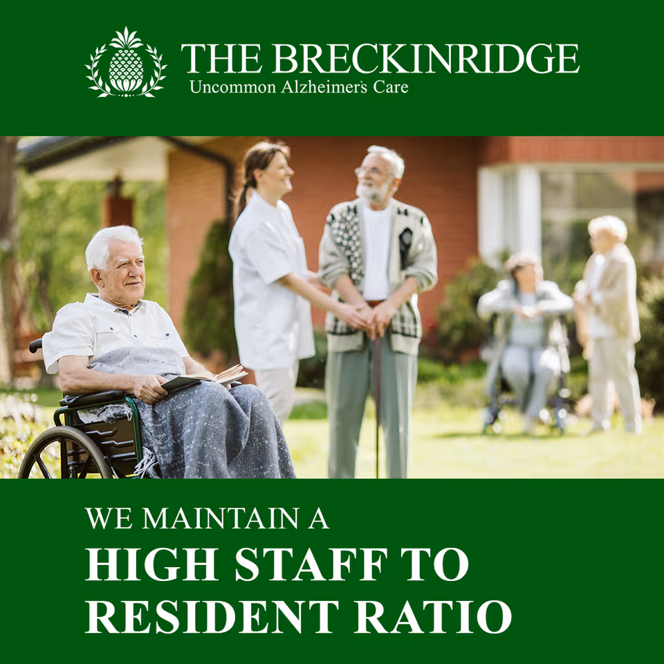 An elderly man in a wheelchair holding a book is outdoors with a caregiver and other elderly individuals in the background, enjoying a sunny day in a garden area of a memory care facility.