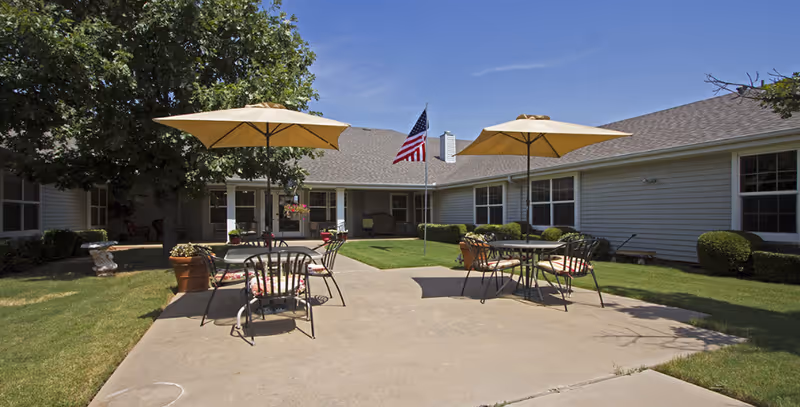 Outdoor patio area at Arbor house with two round tables, each shaded by a large beige umbrella. Each table is surrounded by metal chairs with cushions. The patio is surrounded by a well-maintained lawn, bushes, and a tree. An American flag is mounted on a pole in the center of the lawn, and the building with gray siding and multiple windows forms a U-shape around the patio.