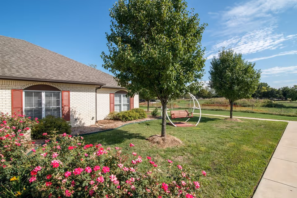Exterior view of a senior living facility building with beige brick walls and red window shutters. The foreground features a garden with blooming pink flowers, green grass, and two trees. A white metal-framed swing with a wooden seat is positioned between the trees. A concrete walkway runs along the right side of the image under a blue sky with scattered clouds.