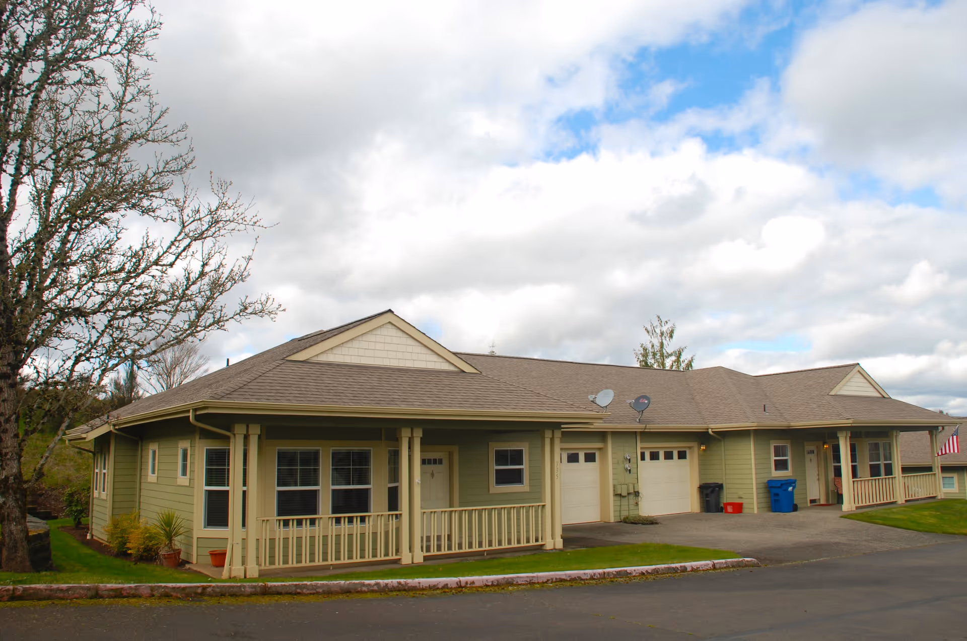 Exterior view of a single-story residential building with green siding, multiple windows, two garage doors, and a small front porch with railings. The building is surrounded by a paved driveway and some grass, with a tree on the left side and a partly cloudy sky overhead.