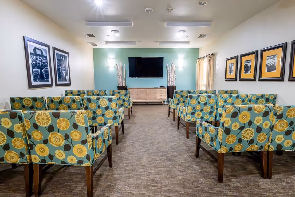 A small theater or media room with rows of armchairs upholstered in a floral pattern facing a wall-mounted flat-screen TV. The walls are decorated with framed black-and-white photos on the left and framed posters with words like 'FEATURE' and 'MOVIES' on the right. The room has carpeted flooring and soft lighting.