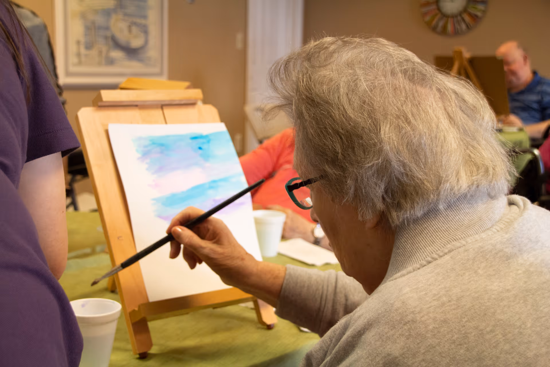 An elderly person with gray hair and glasses is painting on a canvas set on a wooden easel. The painting appears to be an abstract watercolor with shades of blue and purple. Other people are visible in the background, also engaged in painting activities in a cozy indoor setting.