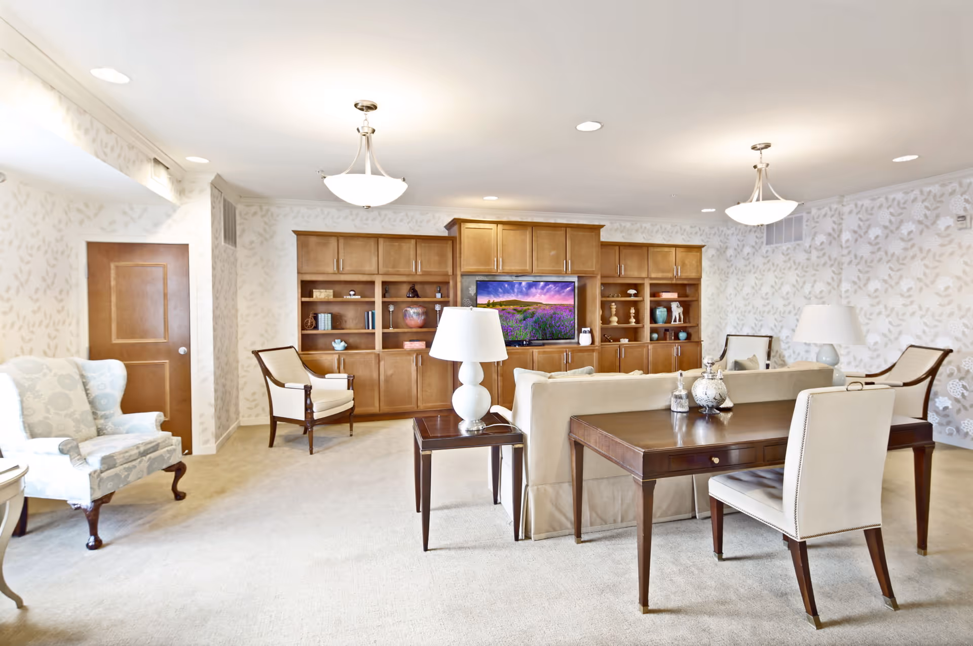 A bright and spacious living room with beige carpet and wallpaper featuring a subtle leaf pattern. The room includes a large wooden entertainment center with a flat-screen TV displaying a scenic landscape, two armchairs, a beige sofa, a wooden side table with a white lamp, and a wooden desk with a chair. The ceiling has recessed lighting and two hanging light fixtures.