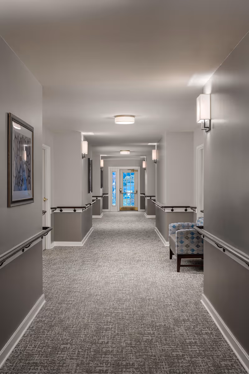 A well-lit, carpeted hallway in a senior living facility with handrails on both sides, light gray walls, framed artwork, and a patterned armchair near the right wall. The hallway leads to a glass door at the far end.