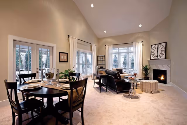 A spacious and well-lit living and dining area featuring a round dining table set for four with plates and glasses, a cozy seating area with a black sofa, a coffee table, a plaid ottoman, a fireplace with a large clock above it, and large windows with white curtains allowing natural light to fill the room.