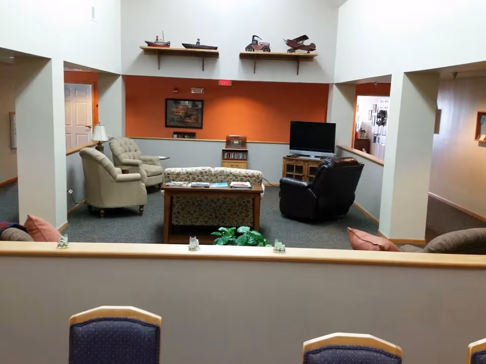 A cozy living room area with a floral patterned sofa, two beige armchairs, and a black recliner arranged around a wooden coffee table. A flat-screen TV sits on a wooden stand against an orange accent wall. Decorative model boats and cars are displayed on a shelf above the seating area. The room has high ceilings and is carpeted, with a few cushions visible on the foreground seating.