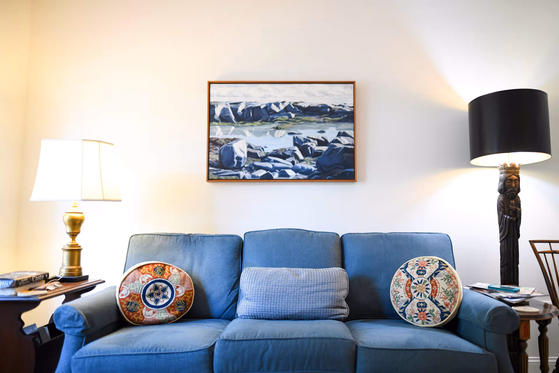 Blue upholstered sofa with patterned round pillows beneath a framed landscape painting, flanked by two table lamps in a living room.