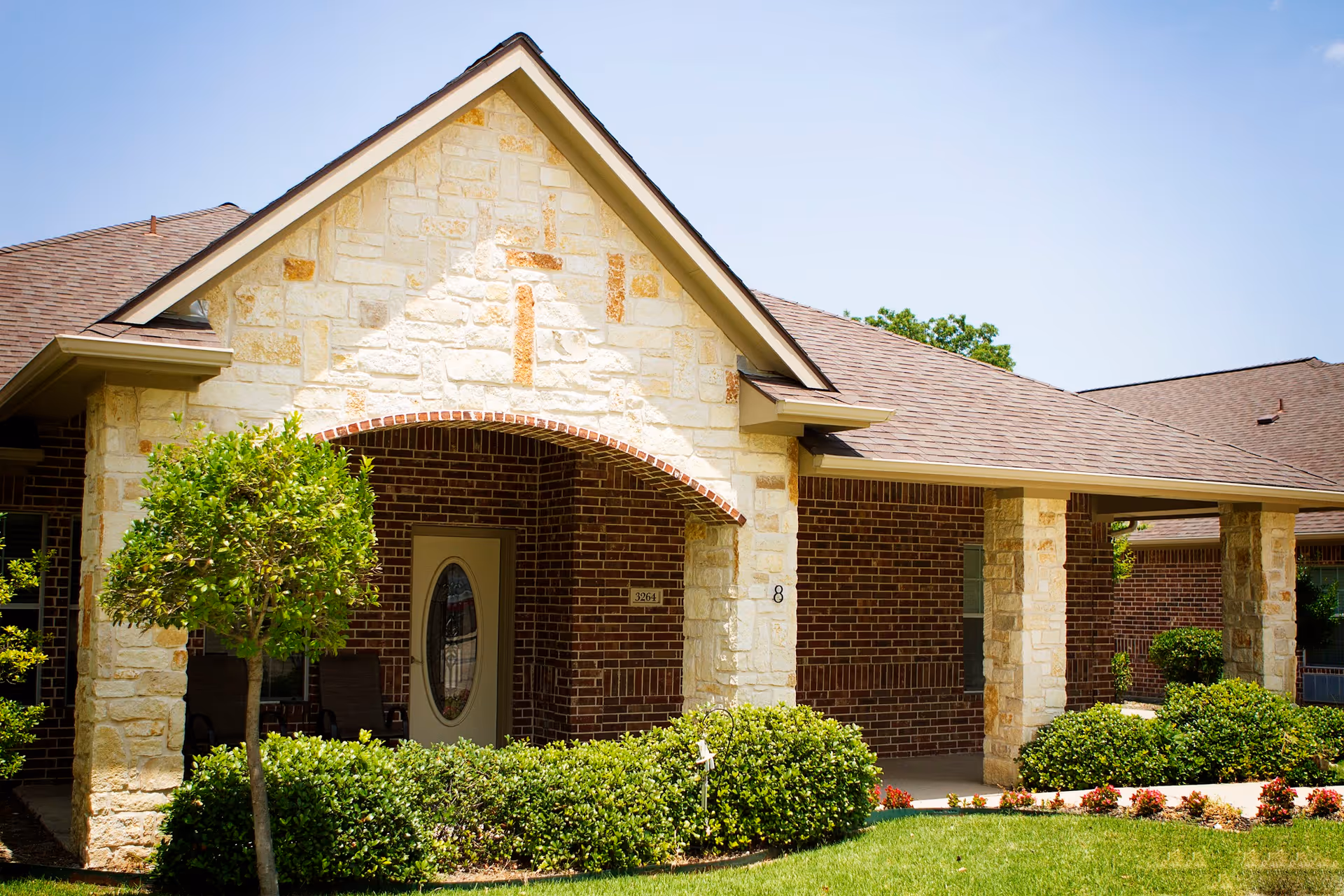 Front entrance of a single-story brick and stone assisted living home with a covered porch, shrubs, and a small tree.