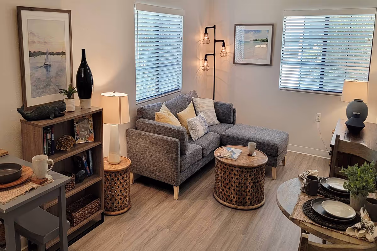 Cozy living room with a gray sectional sofa, round wooden coffee tables, bookshelf, and dining table set near two windows with blinds.